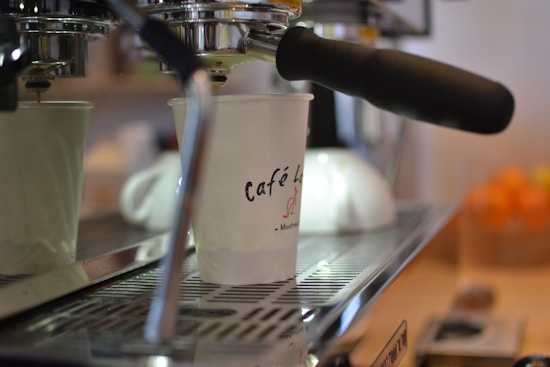 An espresso machine dispensing coffee into a paper cup labeled 'Café' with an indistinct logo. The machine's metal surface has a sleek, polished appearance. The background is softly blurred, with a counter and a bowl of oranges slightly visible.