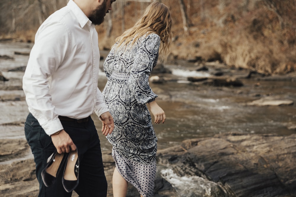 woman near river with man holding her pair of shoes during daytime