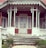 A rustic wooden porch with decorative trim and weathered white paint, featuring a set of stairs leading to an old-fashioned glass-paneled door. The porch has intricate details in the woodwork and is surrounded by small plants and shrubbery.