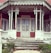 A rustic wooden porch with decorative trim and weathered white paint, featuring a set of stairs leading to an old-fashioned glass-paneled door. The porch has intricate details in the woodwork and is surrounded by small plants and shrubbery.