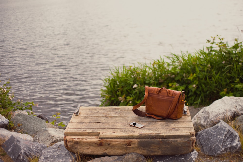 Bolso bandolera de cuero marrón sobre panel de madera marrón cerca del agua
