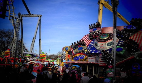 A lively carnival atmosphere with colorful rides and attractions under a bright blue sky. The scene features a star-spangled ride with spinning sections, surrounded by various booths and signs, including one that reads 'Jumper Jumper'. People are visible enjoying the festivities.