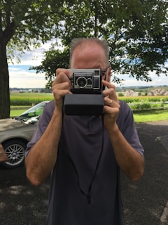 A person is holding an old-fashioned camera, possibly a polaroid, in front of their face. The background features trees and a rural landscape with houses and fields under a partly cloudy sky.