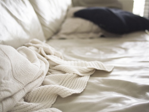 Soft focus image of a bed with a cream-colored knit blanket casually draped over it. A dark pillow is placed in the background on a light bedspread, with natural light coming in from the side creating gentle shadows and highlights.