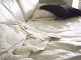 Soft focus image of a bed with a cream-colored knit blanket casually draped over it. A dark pillow is placed in the background on a light bedspread, with natural light coming in from the side creating gentle shadows and highlights.