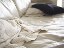 Soft focus image of a bed with a cream-colored knit blanket casually draped over it. A dark pillow is placed in the background on a light bedspread, with natural light coming in from the side creating gentle shadows and highlights.