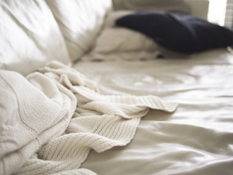 Soft focus image of a bed with a cream-colored knit blanket casually draped over it. A dark pillow is placed in the background on a light bedspread, with natural light coming in from the side creating gentle shadows and highlights.
