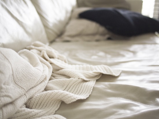 Soft focus image of a bed with a cream-colored knit blanket casually draped over it. A dark pillow is placed in the background on a light bedspread, with natural light coming in from the side creating gentle shadows and highlights.