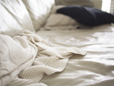 Soft focus image of a bed with a cream-colored knit blanket casually draped over it. A dark pillow is placed in the background on a light bedspread, with natural light coming in from the side creating gentle shadows and highlights.