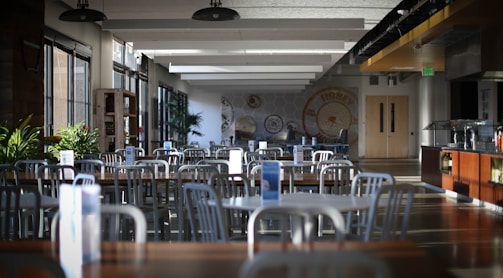 An empty cafeteria with rows of metal chairs and wooden tables. Ambient lighting from large windows creates a calm atmosphere. Potted plants add greenery, and walls are adorned with circular decorative patterns. To the right is a serving counter, and exit doors are visible in the back.