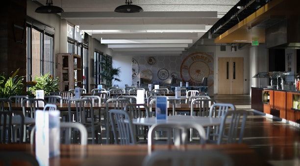 An empty cafeteria with rows of metal chairs and wooden tables. Ambient lighting from large windows creates a calm atmosphere. Potted plants add greenery, and walls are adorned with circular decorative patterns. To the right is a serving counter, and exit doors are visible in the back.