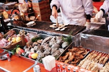 A street food vendor is grilling small round items on a metal grate. The setup includes trays filled with various seafood, meat skewers, and grilled chicken wings. On the left, there's a teapot and assorted condiments, including bottles and decorative lemons and tomatoes. The scene is lively and bustling, reflecting a busy market environment.