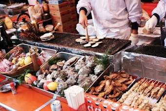 A street food vendor is grilling small round items on a metal grate. The setup includes trays filled with various seafood, meat skewers, and grilled chicken wings. On the left, there's a teapot and assorted condiments, including bottles and decorative lemons and tomatoes. The scene is lively and bustling, reflecting a busy market environment.