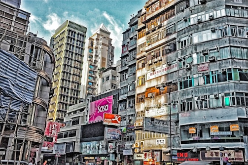 A bustling urban scene with tall buildings densely packed together, showcasing varied architectural styles. Brightly colored advertisements and signs fill the building facades, adding vibrancy. The image captures a cloudy sky above, amidst the tightly knit structures. The atmosphere feels busy and crowded, typical of a city hub.