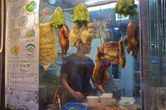 A street food stall displaying various hanging roasted meats including ducks, chickens, and tripe. Fresh lettuce heads are also hanging, and a menu with dishes and prices is visible on the left side. A man, possibly the vendor, stands in the stall wearing a black t-shirt and working with some dishes and noodles on the counter.