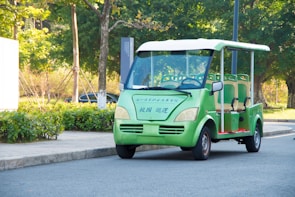 An electric vehicle, similar to a golf cart, is parked on a paved road. The vehicle is green with beige seats and has Chinese characters on its front. Surrounding the vehicle, there is a landscaped area with shrubs, trees, and a paved sidewalk.
