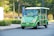 An electric vehicle, similar to a golf cart, is parked on a paved road. The vehicle is green with beige seats and has Chinese characters on its front. Surrounding the vehicle, there is a landscaped area with shrubs, trees, and a paved sidewalk.