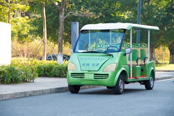 An electric vehicle, similar to a golf cart, is parked on a paved road. The vehicle is green with beige seats and has Chinese characters on its front. Surrounding the vehicle, there is a landscaped area with shrubs, trees, and a paved sidewalk.