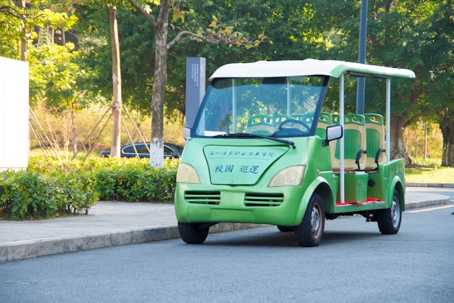 An electric vehicle, similar to a golf cart, is parked on a paved road. The vehicle is green with beige seats and has Chinese characters on its front. Surrounding the vehicle, there is a landscaped area with shrubs, trees, and a paved sidewalk.