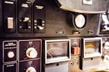 A close-up view of a vintage audio equipment panel with various knobs, switches, and dials. The panel includes controls for tape speed, reverse mode, and microphone and line levels. Several analog meters with needles display sound levels, and there are labels such as VU and REC visible on the dials.