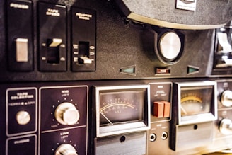 A close-up view of a vintage audio equipment panel with various knobs, switches, and dials. The panel includes controls for tape speed, reverse mode, and microphone and line levels. Several analog meters with needles display sound levels, and there are labels such as VU and REC visible on the dials.
