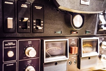 A close-up view of a vintage audio equipment panel with various knobs, switches, and dials. The panel includes controls for tape speed, reverse mode, and microphone and line levels. Several analog meters with needles display sound levels, and there are labels such as VU and REC visible on the dials.