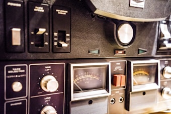 A close-up view of a vintage audio equipment panel with various knobs, switches, and dials. The panel includes controls for tape speed, reverse mode, and microphone and line levels. Several analog meters with needles display sound levels, and there are labels such as VU and REC visible on the dials.