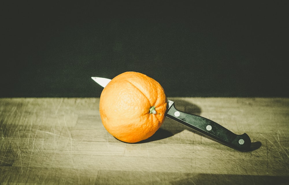 fruta naranja y cuchillo gris y negro sobre tablero de madera marrón fotografía con poca luz