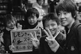 A group of young people stand together, some flashing peace signs, while posing for a photo in a lively setting. A sign in mixed languages is held up by one of the individuals. The scene conveys a sense of camaraderie and playful interaction.