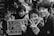 A group of young people stand together, some flashing peace signs, while posing for a photo in a lively setting. A sign in mixed languages is held up by one of the individuals. The scene conveys a sense of camaraderie and playful interaction.