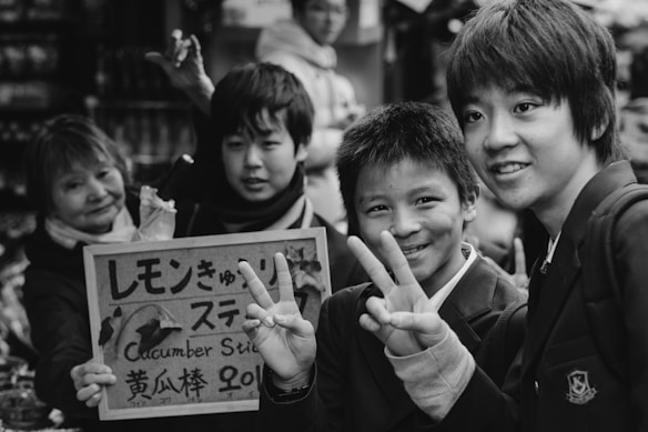 A group of young people stand together, some flashing peace signs, while posing for a photo in a lively setting. A sign in mixed languages is held up by one of the individuals. The scene conveys a sense of camaraderie and playful interaction.