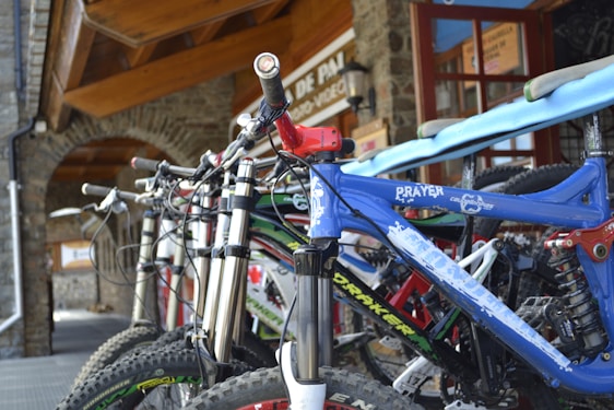 A row of mountain bikes is lined up outside a building with stone walls and a wooden ceiling. The focus is on the nearest bike, which has a blue frame with white text. The building has a sign with visible text in the background.