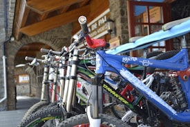 A row of mountain bikes is lined up outside a building with stone walls and a wooden ceiling. The focus is on the nearest bike, which has a blue frame with white text. The building has a sign with visible text in the background.