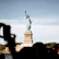 Smiling tourists taking photos near the Statue of Liberty on a bright day.