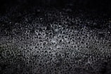A collection of water droplets closely packed together on a dark surface, creating a textured appearance. The droplets vary in size and reflect light, giving them a shiny and glossy look.