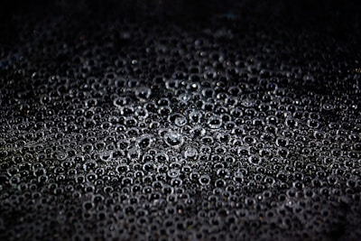 A collection of water droplets closely packed together on a dark surface, creating a textured appearance. The droplets vary in size and reflect light, giving them a shiny and glossy look.