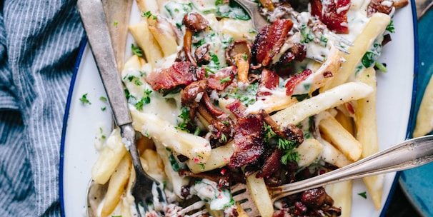 bacon strips and melted cheese topped fries on oval white and blue platter with gray stainless steel forks