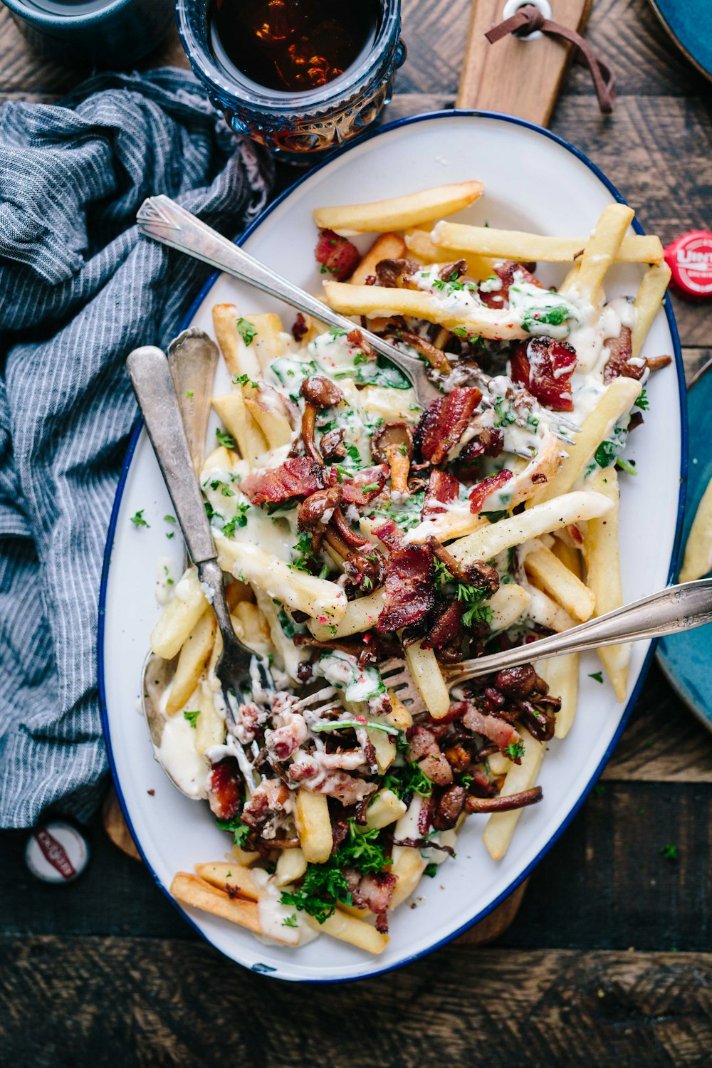 bacon strips and melted cheese topped fries on oval white and blue platter with gray stainless steel forks