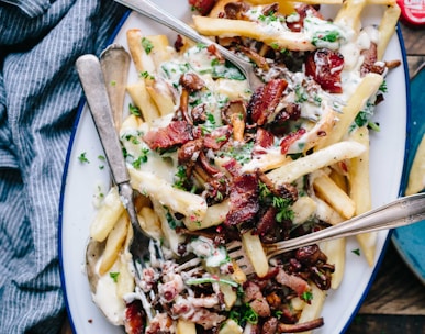 bacon strips and melted cheese topped fries on oval white and blue platter with gray stainless steel forks