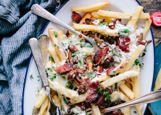 bacon strips and melted cheese topped fries on oval white and blue platter with gray stainless steel forks