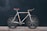 A vintage-style road bicycle with a white frame and brown leather handlebars and seat stands against a dark background. The bike features a minimalist design with thin tires and a single gear mechanism.