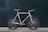 A vintage-style road bicycle with a white frame and brown leather handlebars and seat stands against a dark background. The bike features a minimalist design with thin tires and a single gear mechanism.