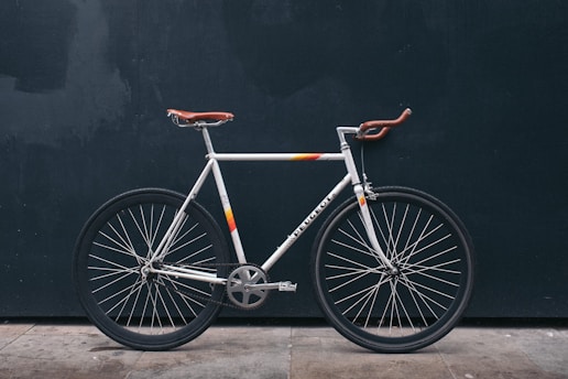 A vintage-style road bicycle with a white frame and brown leather handlebars and seat stands against a dark background. The bike features a minimalist design with thin tires and a single gear mechanism.