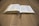 An open book rests on a wooden table, with pages slightly curved. A black ribbon bookmark is placed in the center, and the pages appear dense with text.