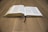 An open book rests on a wooden table, with pages slightly curved. A black ribbon bookmark is placed in the center, and the pages appear dense with text.