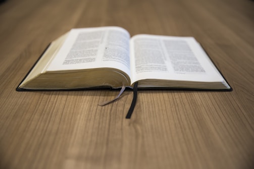 An open book rests on a wooden table, with pages slightly curved. A black ribbon bookmark is placed in the center, and the pages appear dense with text.