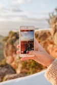 A hand holding a smartphone capturing a photo of an outdoor landscape with rocky terrain blurred in the background. The screen of the phone displays a composition of sky and land under warm lighting.