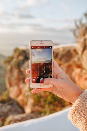 A hand holding a smartphone capturing a photo of an outdoor landscape with rocky terrain blurred in the background. The screen of the phone displays a composition of sky and land under warm lighting.