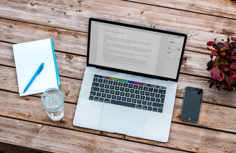 A laptop displaying a document sits on a wooden table accompanied by a closed notebook with a blue pen on top, a glass of water, and a smartphone. A small plant with red leaves is also present, adding a touch of greenery.