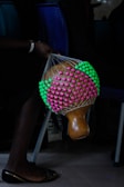 A hand is holding a traditional gourd shaker adorned with a colorful net made of pink and green beads. The background includes chairs, and the scene appears to capture a moment in a dark, indoor setting.